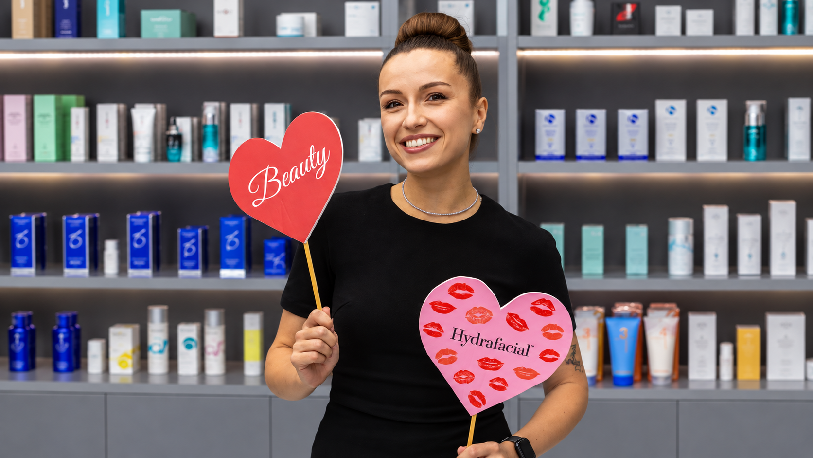 Smiling woman holding Beauty and Hydrafacial heart-shaped signs in front of skincare shelves